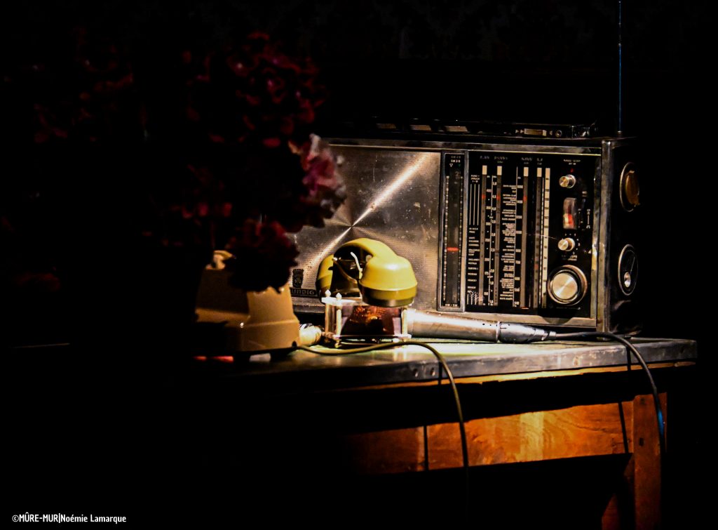 Photo en couleur, lumière tamisée. Scénographie/ décors d'une pièce de théatre qui met en avant un vieux téléphone et une vieille radio sur une table en bois.